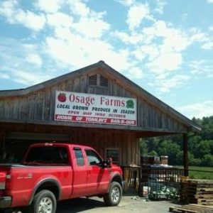 Rustic wooden farm stand with an Osage Farms sign, a red pickup truck parked in front, and pallets nearby.