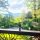 A bottle of red wine and a filled wine glass on a railing, with a lush green garden and blue sky in the background.