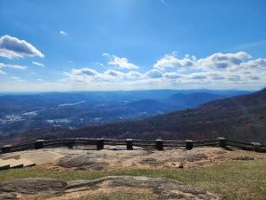View from a mountain overlook: a wooden railing along a rocky viewing platform with distant valleys and layered blue hills under a clear, sunny sky.