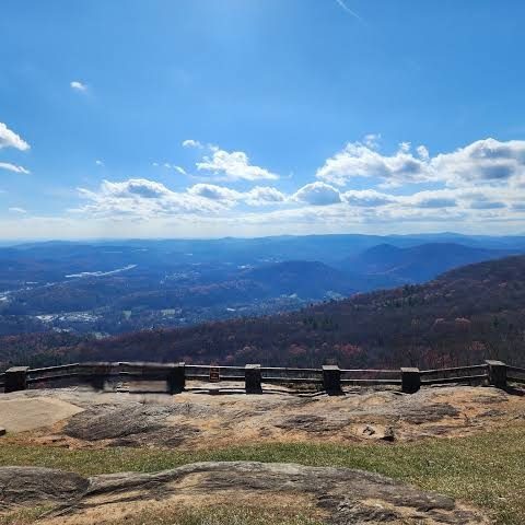 View from a mountain overlook: a wooden railing along a rocky viewing platform with distant valleys and layered blue hills under a clear, sunny sky.