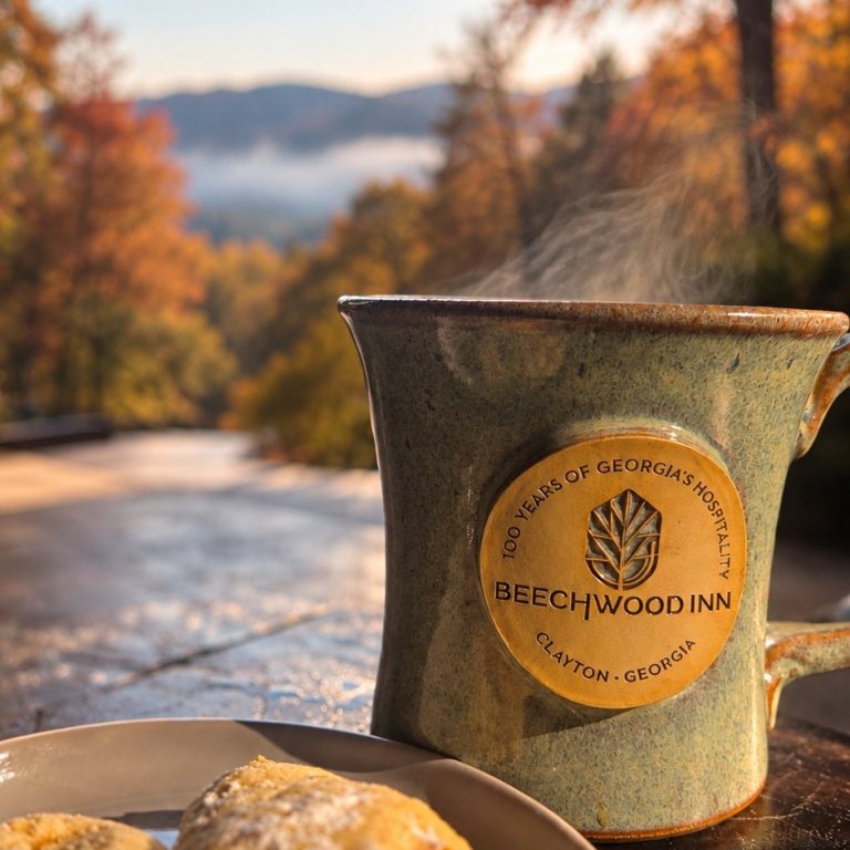 Ceramic mug with Beechwood Inn logo on a wooden table, a plate of scones in the foreground, autumn trees and mountains in the background.