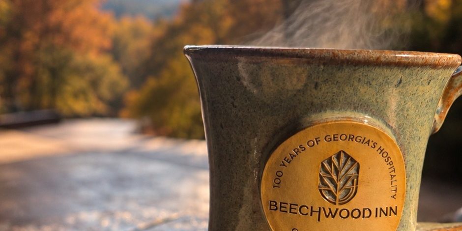 Ceramic mug with Beechwood Inn logo on a wooden table, a plate of scones in the foreground, autumn trees and mountains in the background.