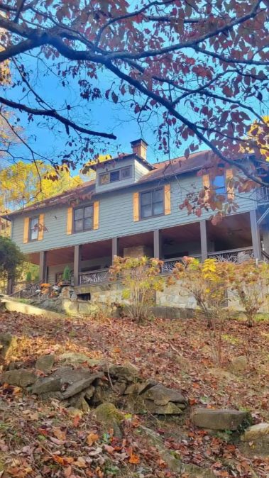 Two-story blue house with yellow shutters on a stone porch, set among autumn trees and fallen leaves on a rocky slope.