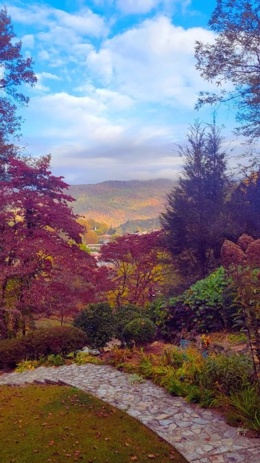 Autumn garden path winding through colorful trees with red, orange, and yellow foliage and distant hills under a blue sky.