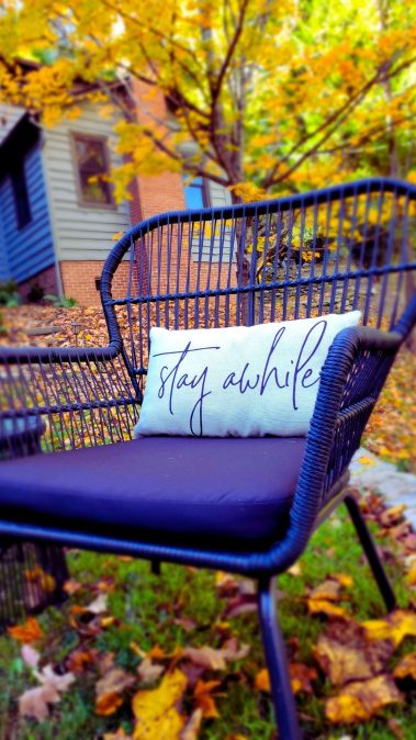 Blue outdoor wicker chair with a light blue cushion that reads 'Stay awhile' in cursive, autumn leaves on the ground and a house in the background.