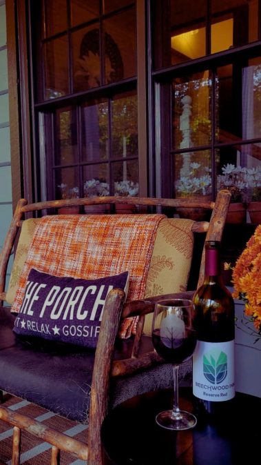 Cozy porch scene with a wooden chair, orange plaid blanket, and a dark cushion reading 'THE PORCH' beside a wine bottle and glass on a dark table.
