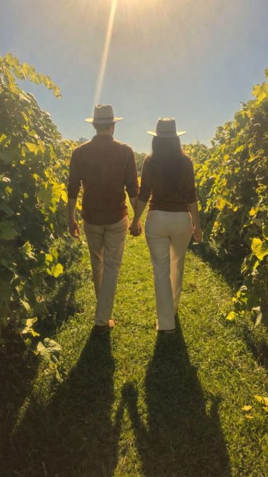 Two people walking hand in hand down a sunlit vineyard path, wearing brown tops and hats, surrounded by green vines.