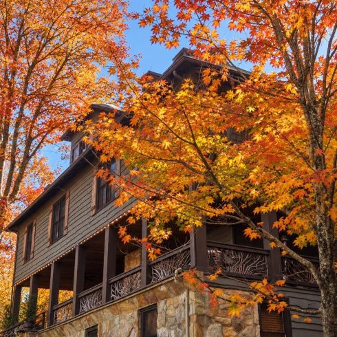 Stone-and-wood house with a wraparound porch, framed by vivid orange autumn leaves against a clear blue sky.