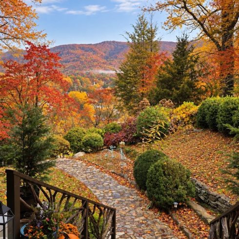 Autumn backyard scene: pumpkins on a wooden porch overlook a stone path, colorful fall foliage, and distant mountains under a blue sky.