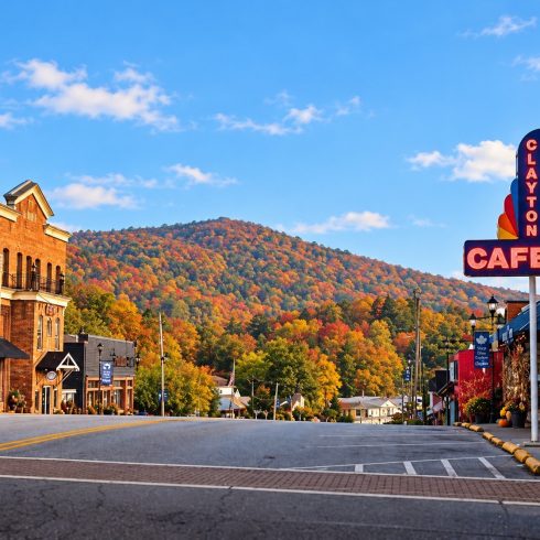 Small-town street with brick buildings, storefronts, and fall-colored trees on a hill under a bright blue sky; a prominent neon 'CAFE' sign and vertical 'Clayton' sign on the right.