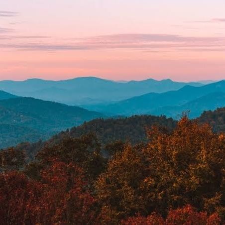 Autumn forest in the foreground with layered blue mountains at sunset.