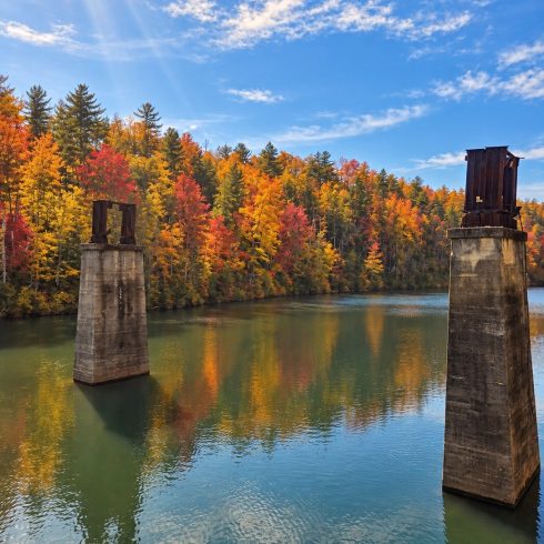 Calm lake reflecting vibrant autumn trees and two rusted concrete pillars rising from the water under a blue sky with sun rays.